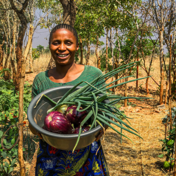 Emelda smiling in a field, holding a basket of fruits and vegetables